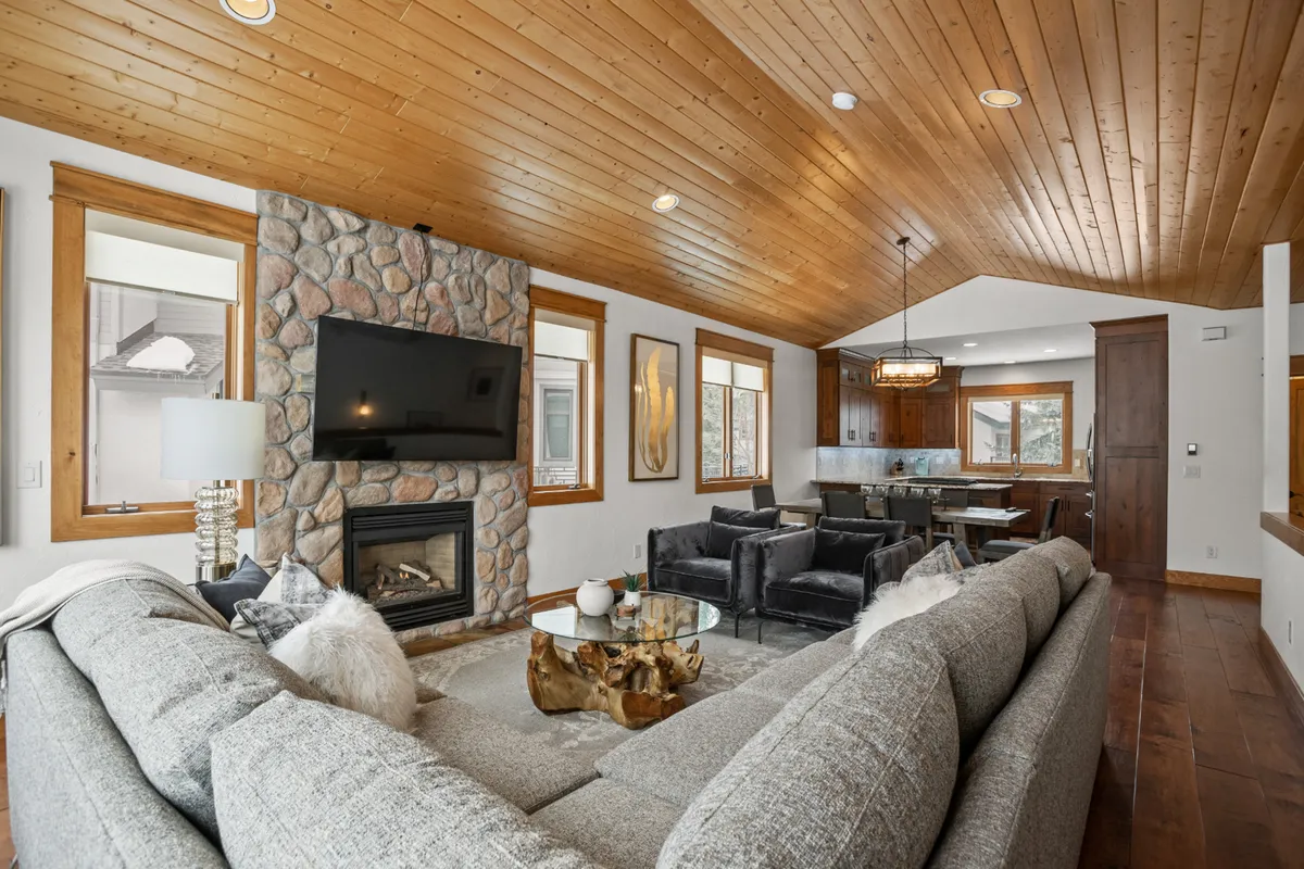 An inviting family living space featuring a stone fireplace, a large gray sofa ideal for group gatherings, artwork on the walls, and a dining area visible in the background. The ambiance is perfect for both cozy winter evenings and summer get-togethers.