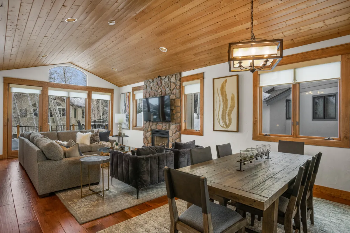 This image features a spacious open-concept living area centered around a striking stone fireplace, with a large sectional sofa and a dining table visible. The space is bathed in natural light, creating an inviting atmosphere ideal for family gatherings and cozy evenings by the fire.
