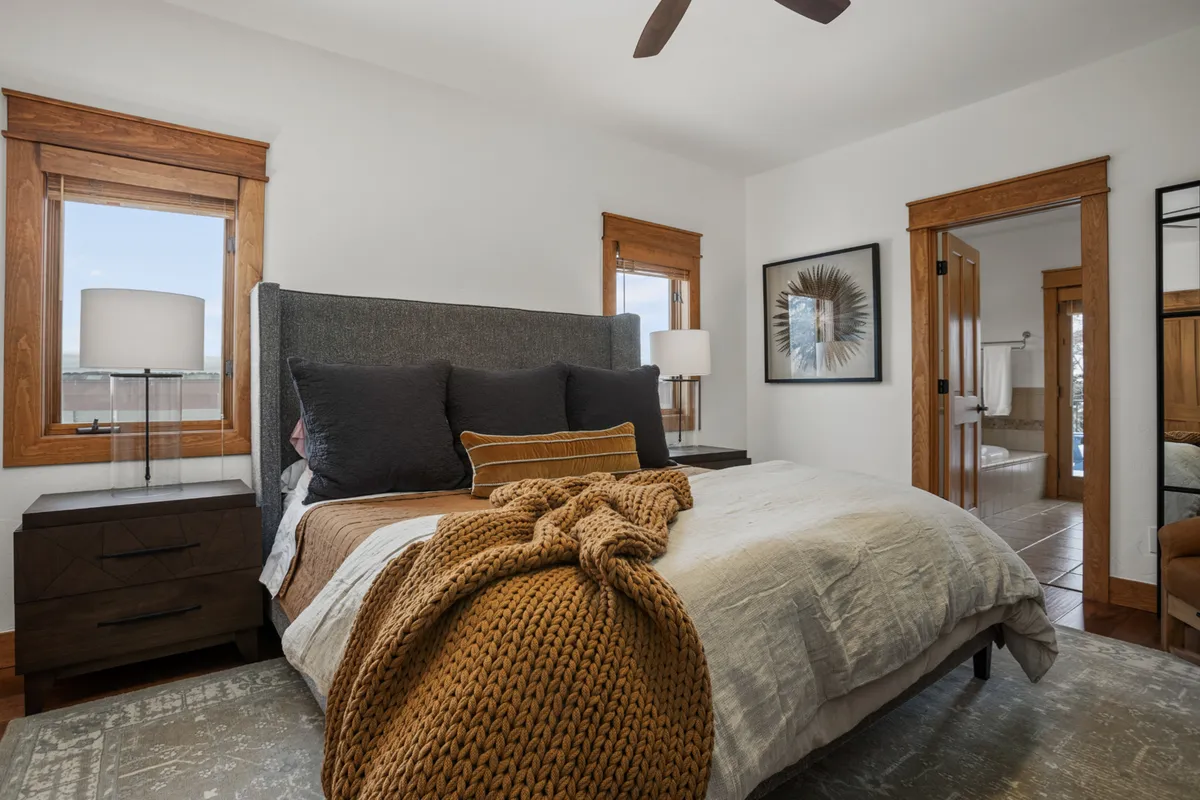 This image showcases a cozy bedroom featuring a bed with a dark gray headboard, a knitted throw blanket, a nightstand with a lamp, and artwork above the bed. Soft, natural light filters into the room, creating a serene and restful atmosphere.