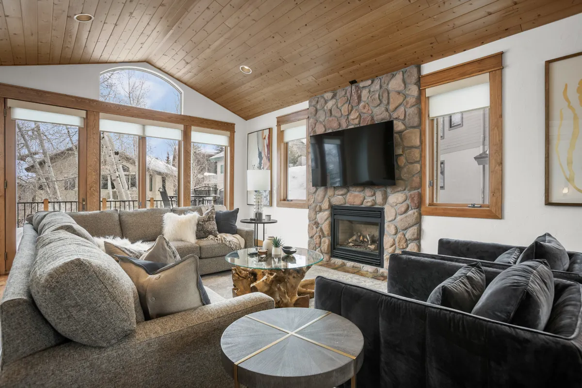 A welcoming living area with a large stone fireplace as the centerpiece, flanked by comfortable seating and bathed in natural light from expansive windows. The cozy space is perfect for socializing, relaxing, or enjoying mountain views through the windows.