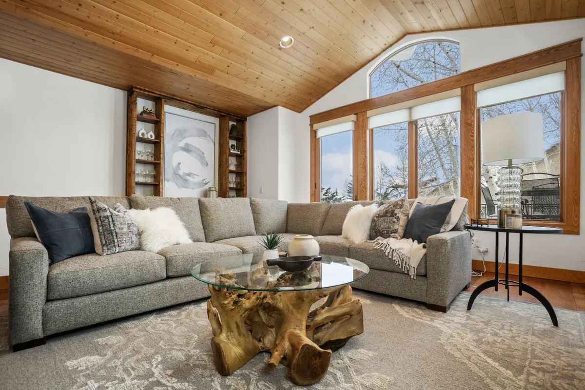 The image showcases a warmly lit living room with a spacious, plush sectional sofa perfect for gathering, a rustic wooden coffee table, and expansive windows that flood the space with natural light, creating a cozy, inviting atmosphere ideal for relaxing or entertaining after a day on the slopes.