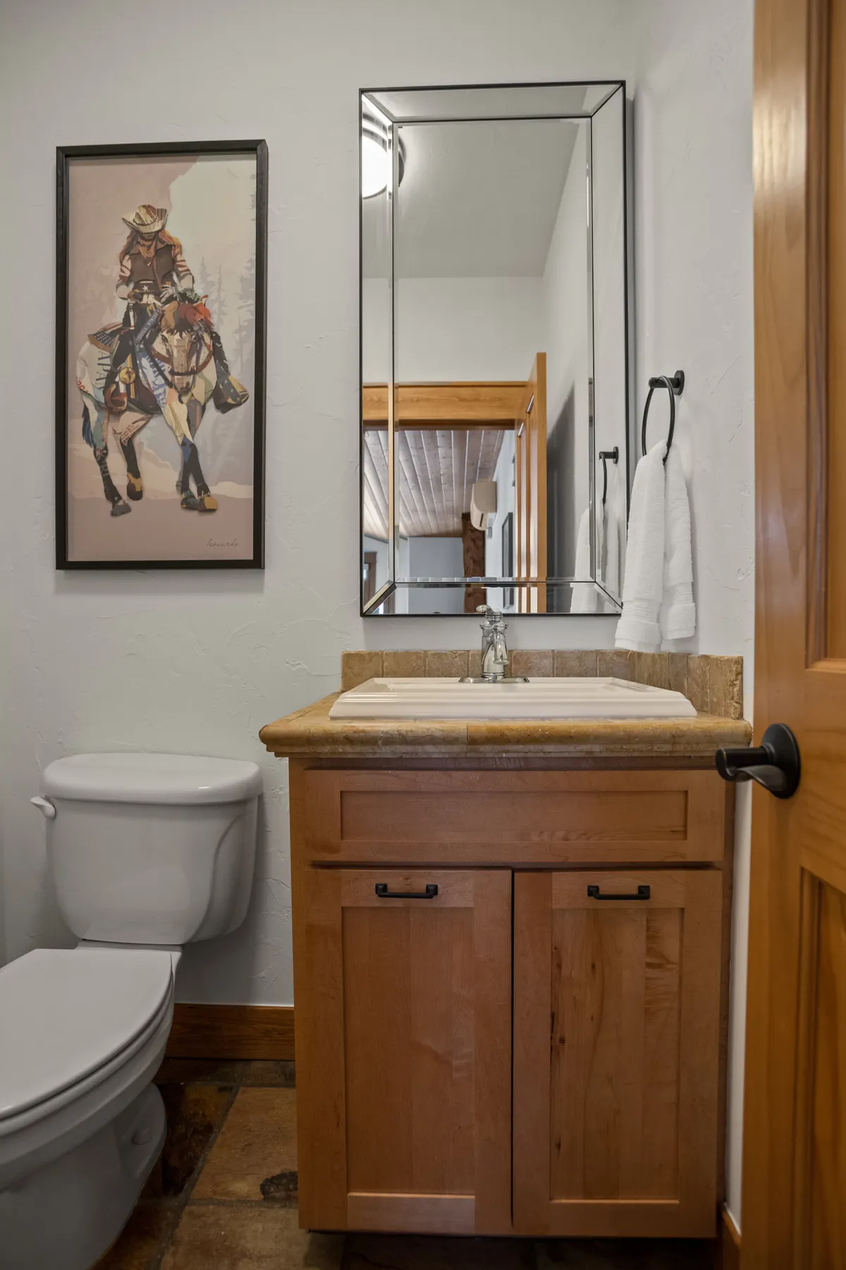 A modern bathroom with a natural stone countertop, modern sink, and wooden cabinetry, highlighted by a framed artwork of a cowboy on horseback, creating a welcoming and subtly adventurous tone.