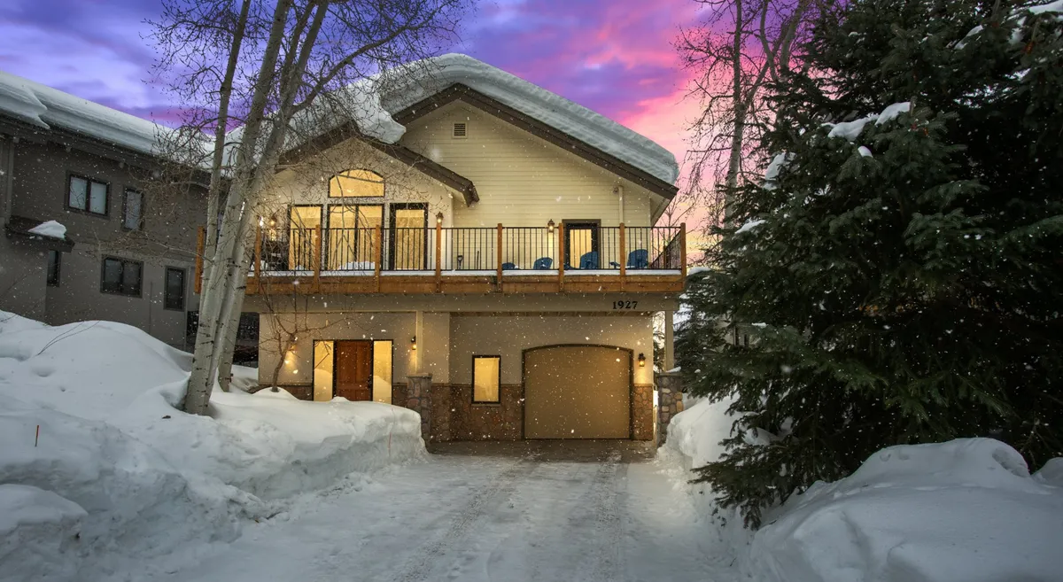 This exterior shot showcases the home's large balcony, the inviting glow from the windows, and a snow-blanketed driveway. The scene suggests cozy evenings after a day on the slopes and hints at après-ski gatherings, stargazing, and relaxing in a private mountain retreat.