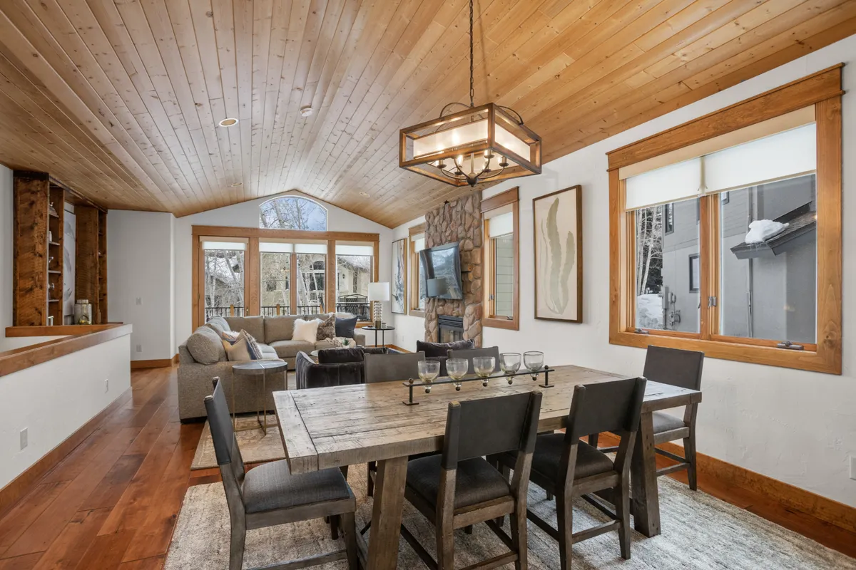 A combined dining and living area with a prominent stone fireplace, stylish chandelier, large windows, and plush sofa seating. The setup is ideal for both formal dinners and relaxed conversations, with cozy fireside ambiance.