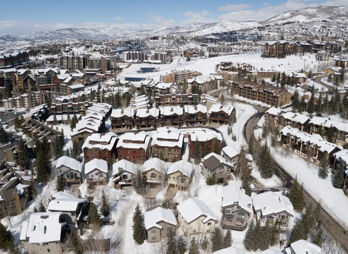 A scenic view of a residential neighborhood with snow-covered rooftops, a winding road, and a stunning mountain backdrop. Clusters of homes are nestled along the road, suggesting easy access to outdoor spaces and trails leading toward the mountains.