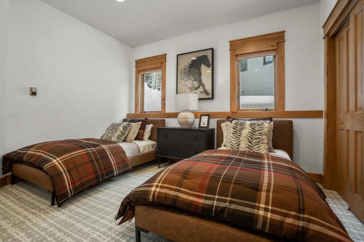 A bedroom with two twin beds dressed in plaid blankets, a black nightstand, wall art, and large windows letting in natural light. The space feels comfortable and thoughtfully arranged, suitable for children, grandparents, or friends.