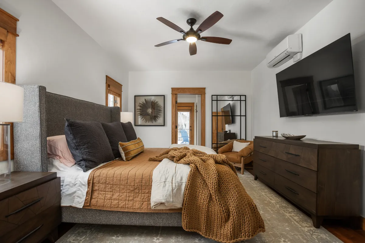 A bedroom with a comfortable bed, decorative mirror, TV atop a dresser, and an armchair. Natural light fills the room, while the layout suggests both relaxation and functionality, ideal for winding down with entertainment or a good book.