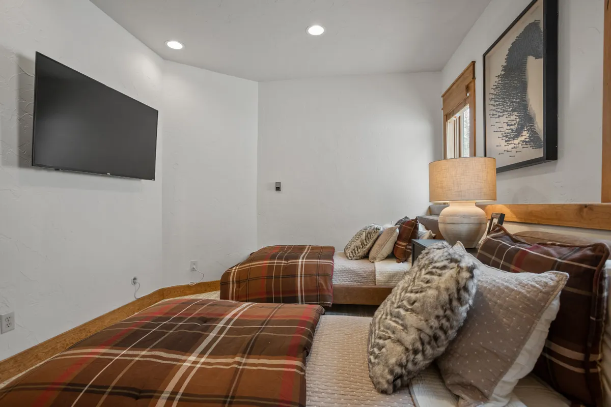 A bedroom featuring plaid bedding, decorative pillows, a wall-mounted TV, a table lamp, and framed artwork. The room is inviting and tailored for relaxation, reading, and enjoying entertainment.