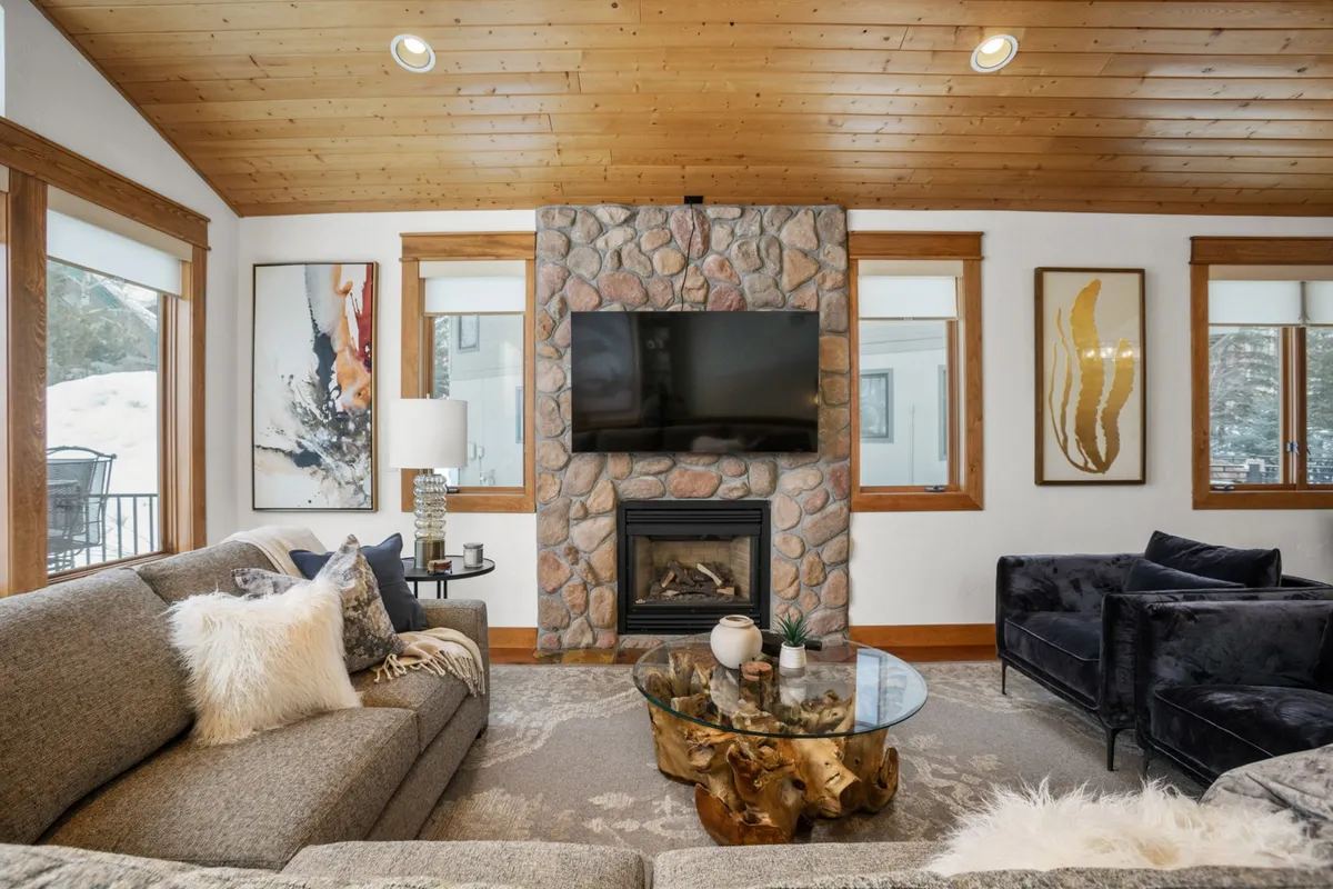 A welcoming living room anchored by a stone fireplace, with a plush couch, a coffee table, and large windows that bathe the space in natural light. Artwork adds warmth, and the setting invites guests to relax, socialize, or enjoy a hot drink after seasonal adventures.